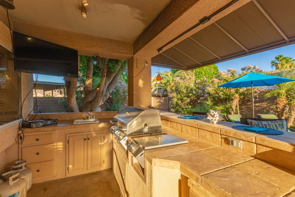 This view features an outdoor kitchen grill with bar seating, decorative elements, and a clear view of the spacious backyard. The setup encourages group cooking, casual barbecues, and outdoor entertaining, with a relaxed and social mood enhanced by the umbrella-shaded seating.