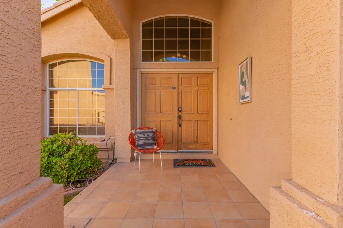 This exterior image features the home's wooden front door, framed by large windows and tile flooring, with a vibrant red chair accenting the space. The scene is bathed in natural light, giving a sense of arrival and welcoming ambiance.