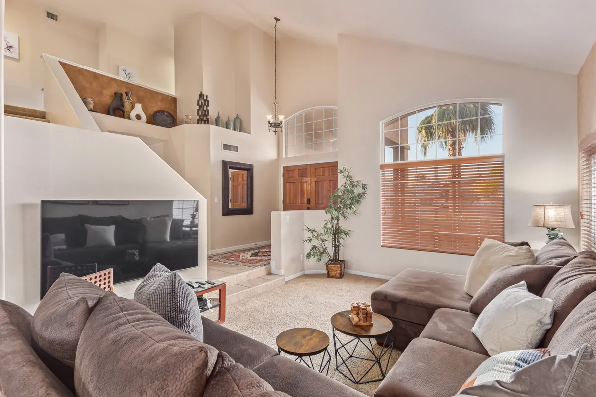 A cozy living room features a large sectional sofa facing a flat-screen TV, with decorative shelves and a window with blinds adding homey touches. The space is ideal for movie nights, casual conversations, or relaxing after a day of entertaining.
