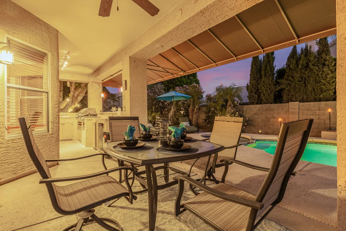 An inviting outdoor living space featuring a dining table set beneath an umbrella, a sparkling swimming pool, and a fully equipped outdoor kitchen. Palm trees and lush landscaping frame the area, while sunset hues illuminate the sky for a true Arizona retreat.