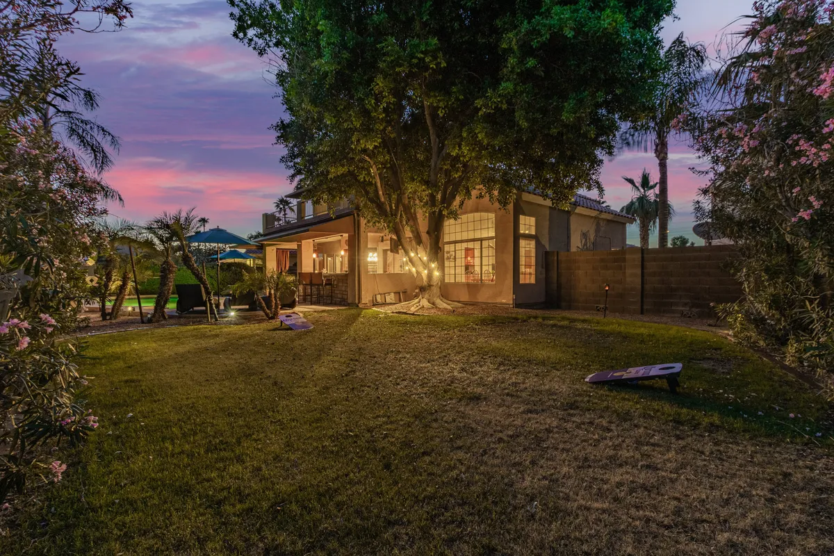A tranquil exterior shot showcasing a large tree shading a well-furnished patio with a dining table and outdoor seating. The area is designed for outdoor dining or relaxed evening gatherings, set against the backdrop of a peaceful yard.