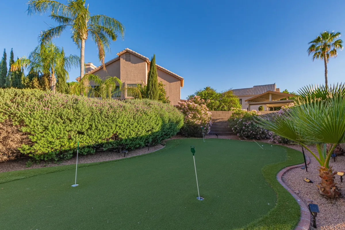 A private putting green surrounded by palm trees, lush greenery, and views of the house's exterior. The scene is tranquil and playful—a dedicated spot for golf practice or friendly competition, complete with sunny skies above.