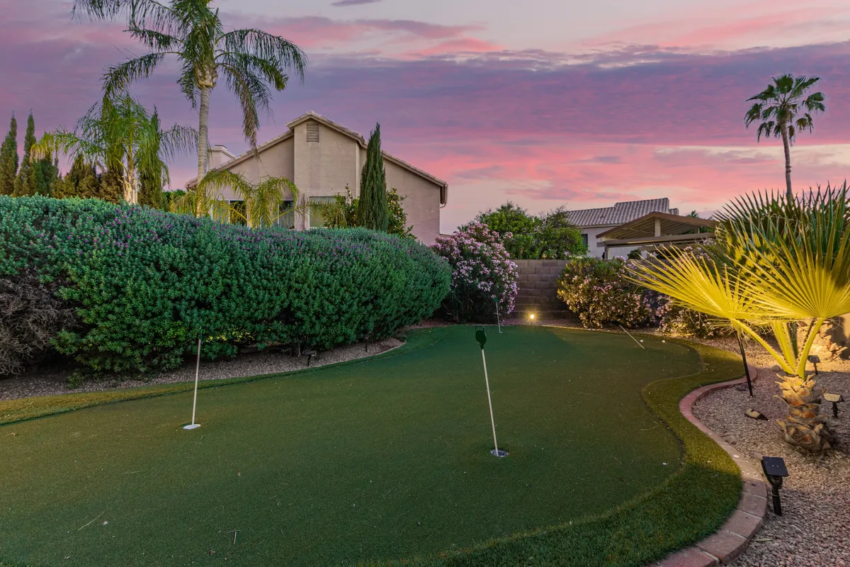 A beautifully landscaped backyard with a vibrant putting green surrounded by lush greenery, colorful flowers, and tall palm trees. The home is visible in the background, and the scene is set for evening fun and family gatherings.