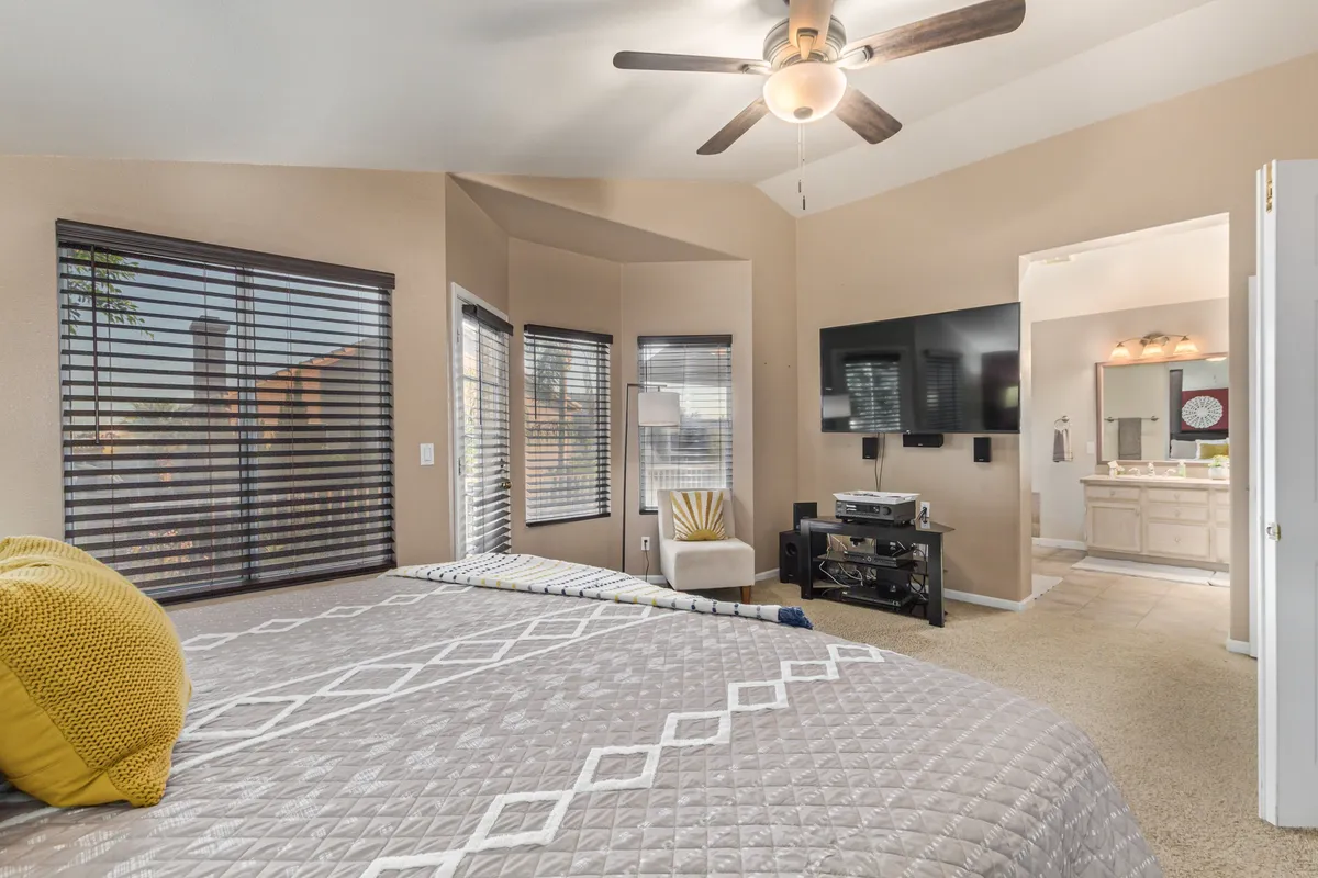 A bright, inviting bedroom features a plush bed with a quilted pattern, large windows with blinds allowing for abundant natural light, a comfortable chair for relaxing, and a TV mounted on the wall. The cozy atmosphere suggests a perfect place for unwinding after a long day.